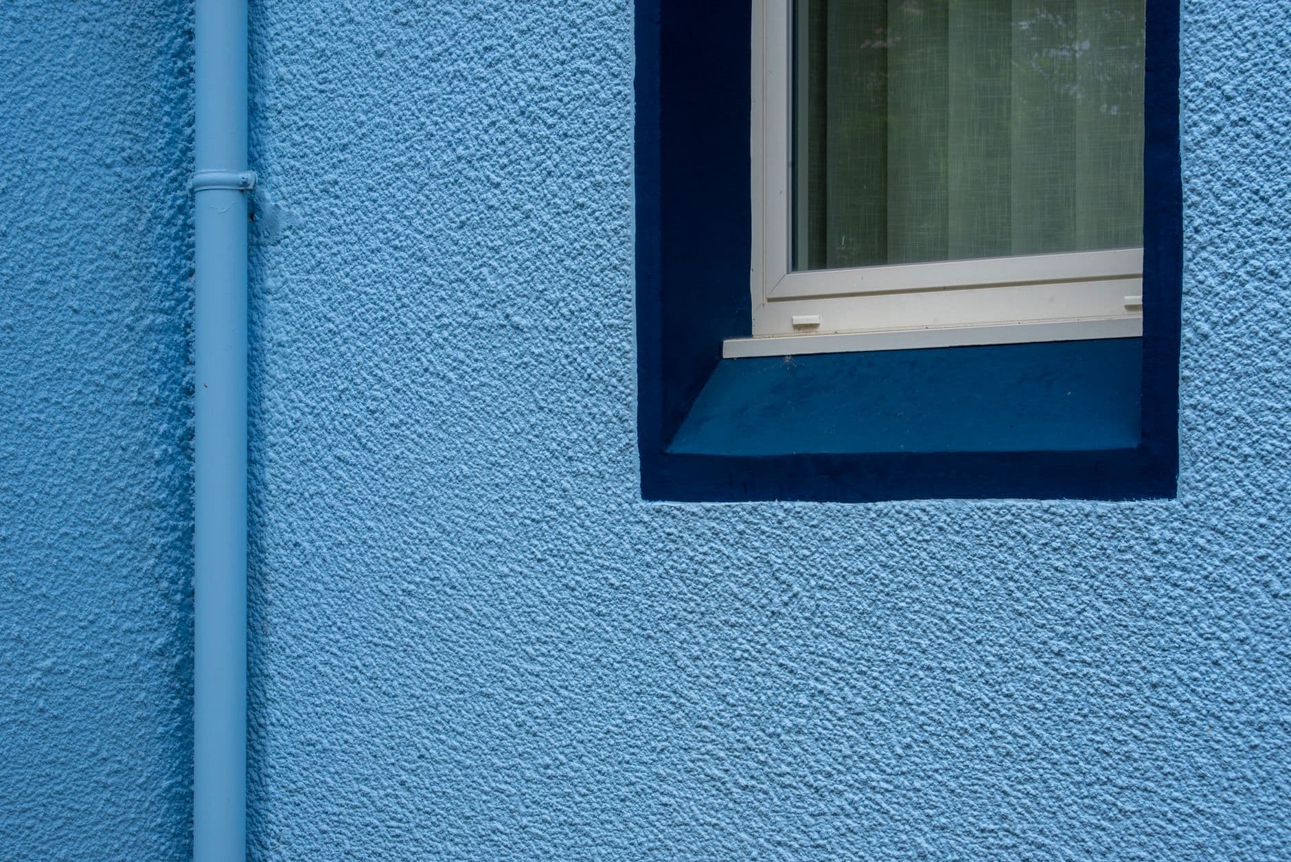 Blauwe buitengevel met korrelige structuurpleister, een raam met donkerblauwe omlijsting en een regenpijp